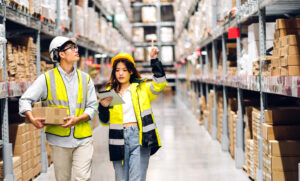 An Asian man and woman in safety vests in a distribution warehouse looking at a tablet and enjoying increased efficiency due to ai automation consulting for distributors.