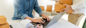 Startup small business entrepreneur SME using an AI tool for small businesses on his laptop while another organize delivery boxes.