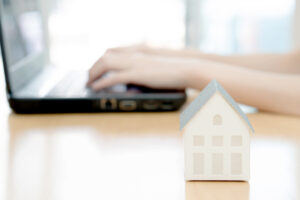 Hand on computer keyboard and mini house in foreground.